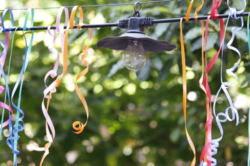 Nature: Light hanging outside on a wire with colored ribbons hanging down with green trees as a background.