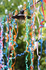 Nature: Light hanging outside on a wire with colored ribbons hanging down with green trees as a background.