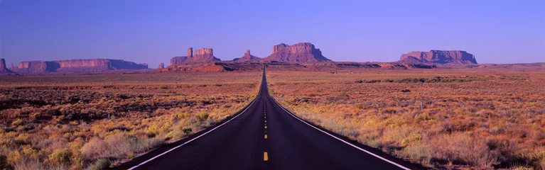 Fotobehang This is Route 163 that runs through the Navajo Indian Reservation. The road runs up the middle and gets smaller into infinity. The red rocks of Monument Valley are in the background. The scrub plants of the desert are on either side of the road. © spiritofamerica