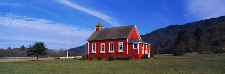 This is the Stone Lagoon School House. It is a one room school house. It is painted a bright red with white trim on the windows. There is a flag pole with no flag on it. The sky is blue and it is surrounded by green grass, bushes and trees. It is located in Northern California, not far from the Oregon border.