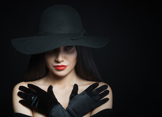 Woman in big black hat with arms across, studio portrait, dark background