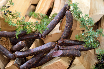 Originale dunkel geräucherte Südtiroler Kaminwurzen vor Holzscheite mit frischem Wacholder platziert 