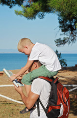 Father giving his son piggyback ride outdoors. Man and boy son looking at map in front of sea, active summer holiday vacation, family travel photo