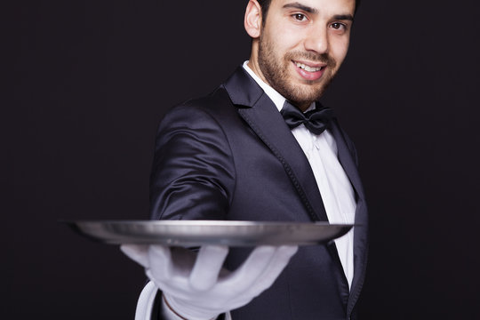 Smiling waiter holding an empty silver tray against dark backgro