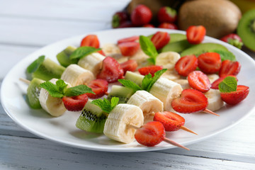 Fresh fruits on skewers in plate on wooden table, closeup