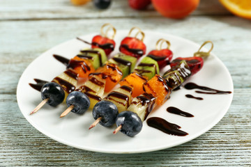 Fresh fruits on skewers in plate on wooden table, closeup