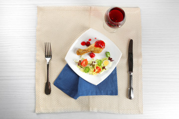 Dish of baked chicken leg and vegetable salad in white plate with glass of wine on table with napkin, top view
