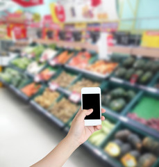 Hand holding mobile phone with Vegetables and fruit on shelf in
