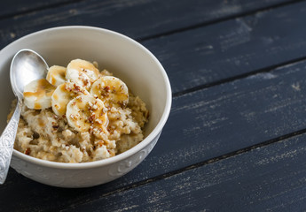 oatmeal with banana, caramel sauce and pecan nuts in a white bowl on a dark wooden surface, a delicious and healthy Breakfast