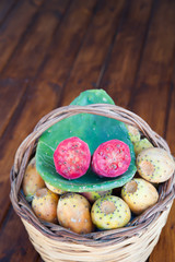 Red cut in half prickly pear on a cactus leave and a wicker basket full of just picked prickly pears on a wooden table