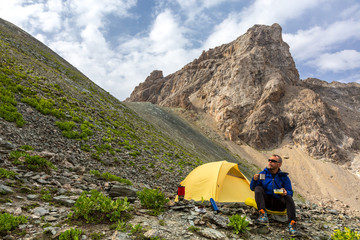 Traveling man eating meal Hiker sitting aside yellow camping tent and having lunch stove and cooking gear mountain landscape on background