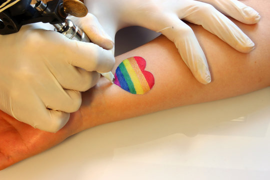 tattooer showing process of making a tattoo on young  woman wrist. Tattoo design on the heart painted in the colors of the rainbow