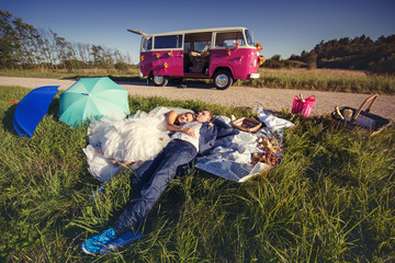 Newlyweds relax on picnic blanket on the wayside, cheerful wedding car in the background.