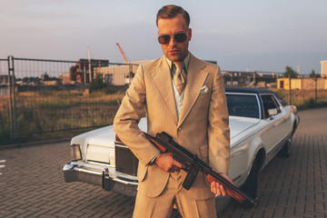 Retro 1970s gangster holding gun standing in front of vintage ca
