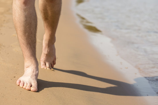 man with hairy legs goes barefoot on the sand beach along the shore near the water with waves