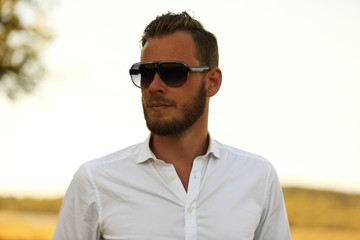 A man wearing a white shirt and dark sunglasses, standing against a large yellow field looking away from camera. A great sunny summer day.