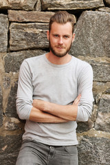 Attractive man wearing a grey sweater standing in front of a wall outside on a sunny summer day. 