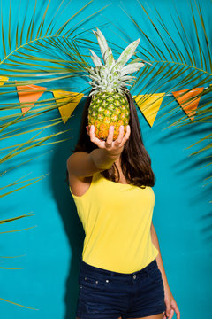 Young woman covering her face with a pineapple. Summer background with palm tree foliage and garland