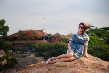 brunette girl in short grey frock sits on rock against villas