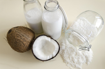 Coconut products. Cracked open coconut with meat cut in half, grounded flakes in a mason jar, flour and fresh milk in glass bottles on a table with red ruby background.