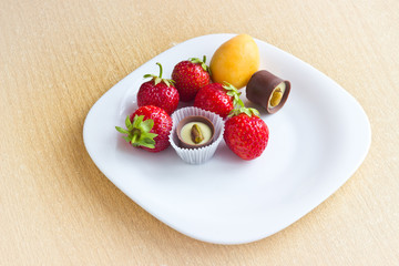 Fresh ripe strawberries, apricot and chocolate candy on white dish on wooden table