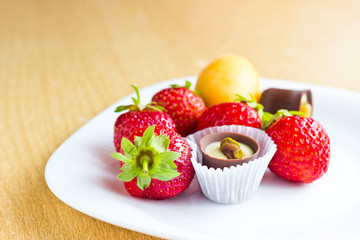 Fresh ripe strawberries, apricot and chocolate candy on white dish on wooden table