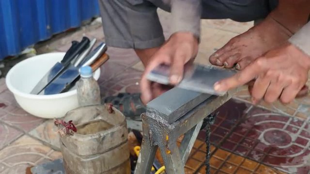 Close-up on a knife sharpener whetting a cleaver on the sidewalk and running his thumb across to check the blade