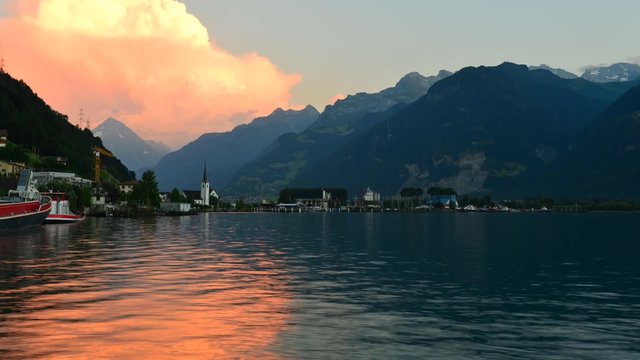 Evening clouds are painted in bright orange color. Mountain landscape.Reflections in the lake. In the distance the small town of in Fluelen in Switzerland. Time Lapse