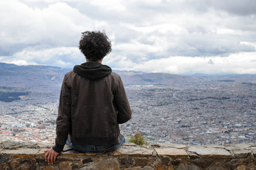Anonymous guy with black curly hair sitting on stone wall overlooking Bogota city 