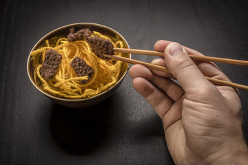 Food knitted from woolen thread lies on a plate, as a Chinese dish of noodles and meat, the human hand holding a wooden spokes like sticks on a dark background