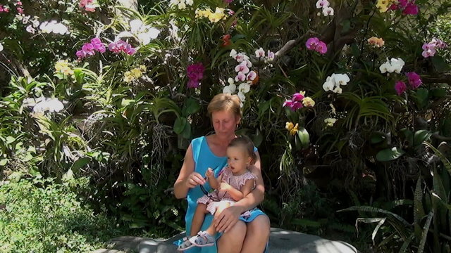 baby sitting on the lap of a woman in a blue dress and playing with sunglasses on the background of trees and beautiful multi-colored orchids
