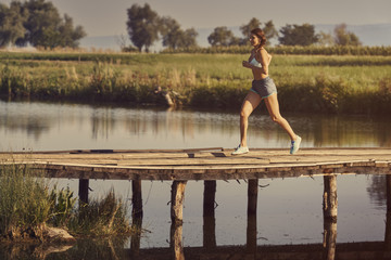 Happy athletic young woman in summer sportswear running, sprinting on a rustic wooden footbridge across a pond at sunrise. Calories burning. Healthy lifestyle. Toned color edit.