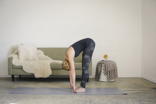 A blonde woman in a black leotard and leggings, standing on a yoga mat in a room, doing yoga, bending down, touching her toes with her hands.