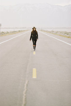 A woman carrying a backpack walking down the centre line of a  country road.