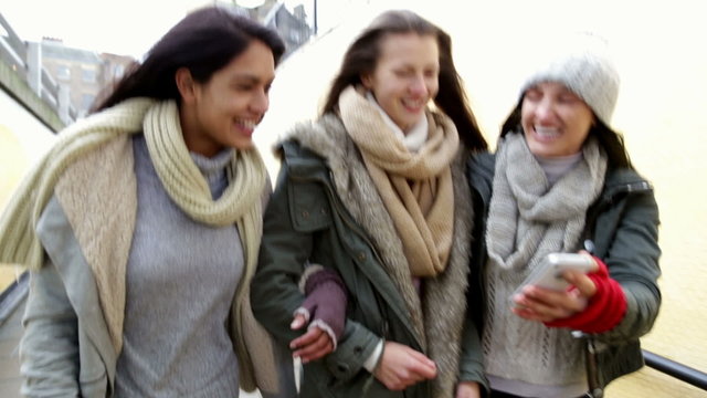 Three young women walking together down towards an underpass with their arms linked, looking at something on a smartphone.