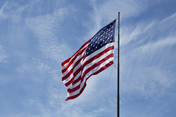 American Flag flutters in the breeze of a crisp clear Spring day. Old Glory is shot against a light blue sky with a few wispy clouds. 
