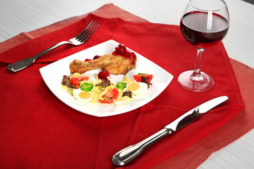 Dish of baked chicken leg and vegetable salad in white plate with glass of wine on table with napkin, closeup