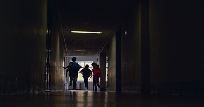 Rear view of elementary students running along dark school corridor 