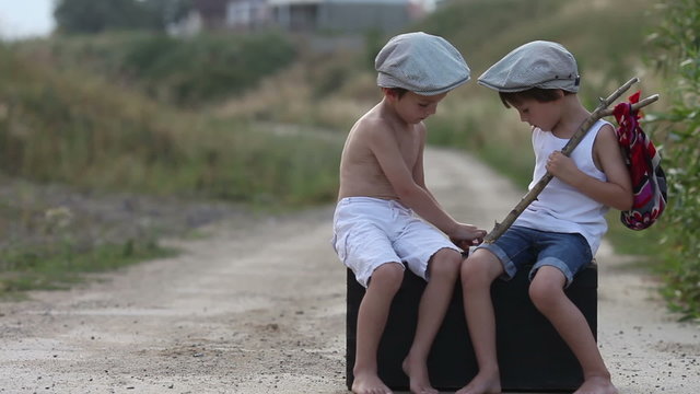 Two boys, sitting on a big old vintage suitcase, playing with toys and eating bread