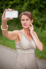 Kopf und Schultern einer Frau die ein elegantes graues Kleid trägt. Sie posing im Freien, während Sie mit ernsten Gesichtsausdruck ein Selfie (Selbstporträt) mit einem intelligenten Handy macht.