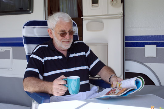 Rentner sitzt Kaffee trinkend  vor seinem Wohnmobil oder Wohnwagen und liest