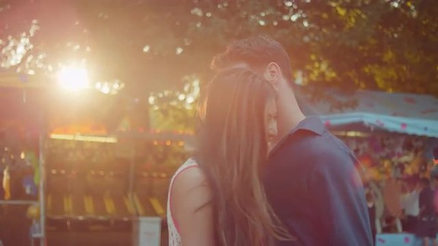 A Couple At A Fair With Their Arms Around Each Other As The Sun Sets Behind Them