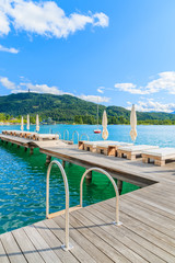 Fototapeta na wymiar Sunchairs on wooden pier and view of beautiful alpine lake Worthersee in summer, Austria