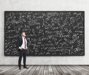 Side view of a confident young man who stands in front of black chalkboard which is full of math...