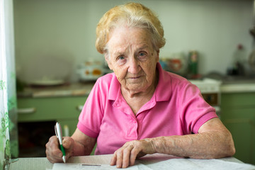 Elderly senior woman populates handle her utility bills notices, sitting at the table in the kitchen.