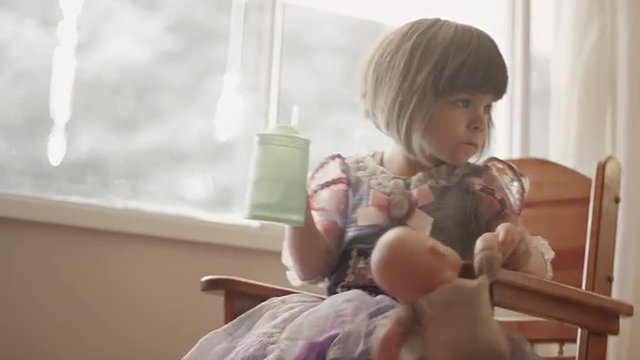 A little girl wearing a princess dress sits by a window and drinks from a sippy cup
