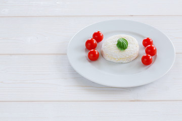 Camembert round entirely with cherry tomatoes and basil leaf on a white plate and a white wooden background