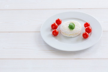 Camembert round entirely with cherry tomatoes and basil leaf on a white plate and a white wooden background