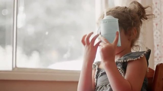 A little girl sitting by a window covers her face with her sippy cup
