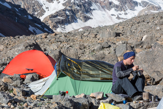 Female Climber Eating Ascetic Breakfast Mountain Expedition Camp Green and Red Tents Woman Sitting and Eating Meal Drinking Tea Steep Rocky and Ice Wall on Background